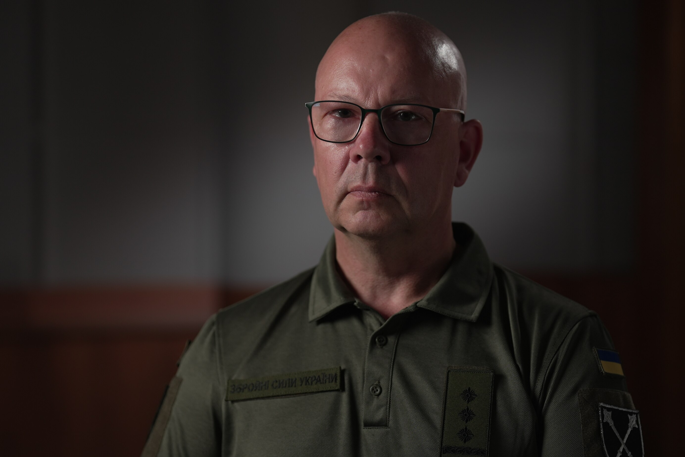 A bald man in a green shirt and glasses looks to the camera
