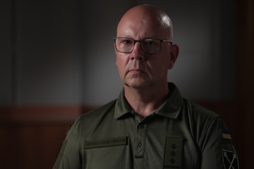 A bald man in a green shirt and glasses looks to the camera