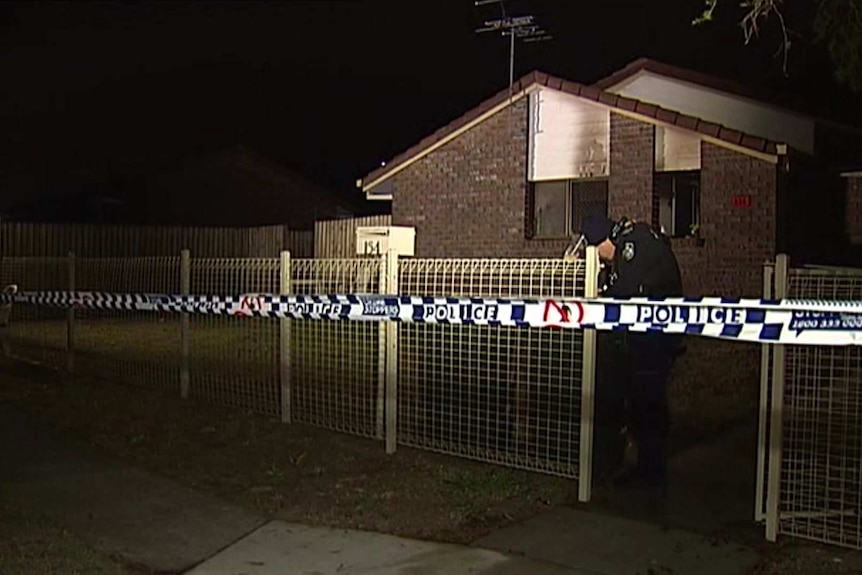 A low-set brick house with a police officer and police tape at the front at night.