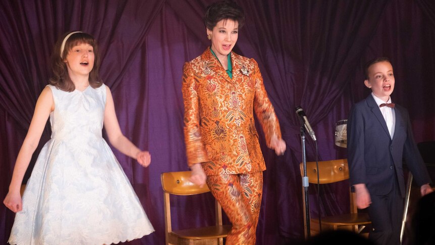 Scene from the movie Judy featuring Renee Zellweger as Judy Garland in orange paisley suit, flanked on stage by her two children