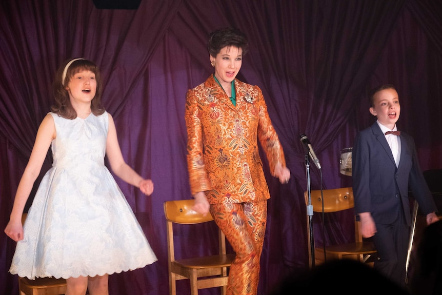 Scene from the movie Judy featuring Renee Zellweger as Judy Garland in orange paisley suit, flanked on stage by her two children