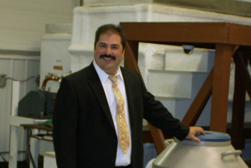 A man in a black suit and yellow tie with a moustache stands with his hand resting on an unseen item