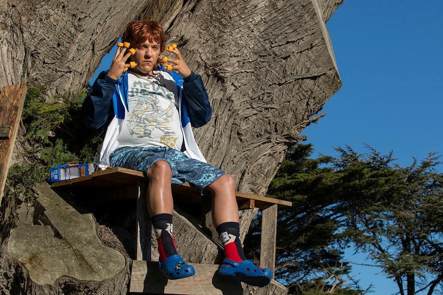 A man dressed as a teenage boy, sits in a tree with cheezles on his fingers.