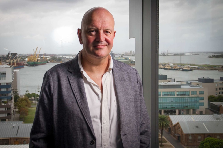 A man in a grey jacket stands in front of a window overlooking an industrial port.