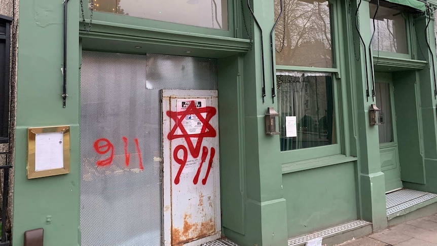 A green cafe shopfront shows a metal door sprayed with a red Star of David and two '9 11' phrases.