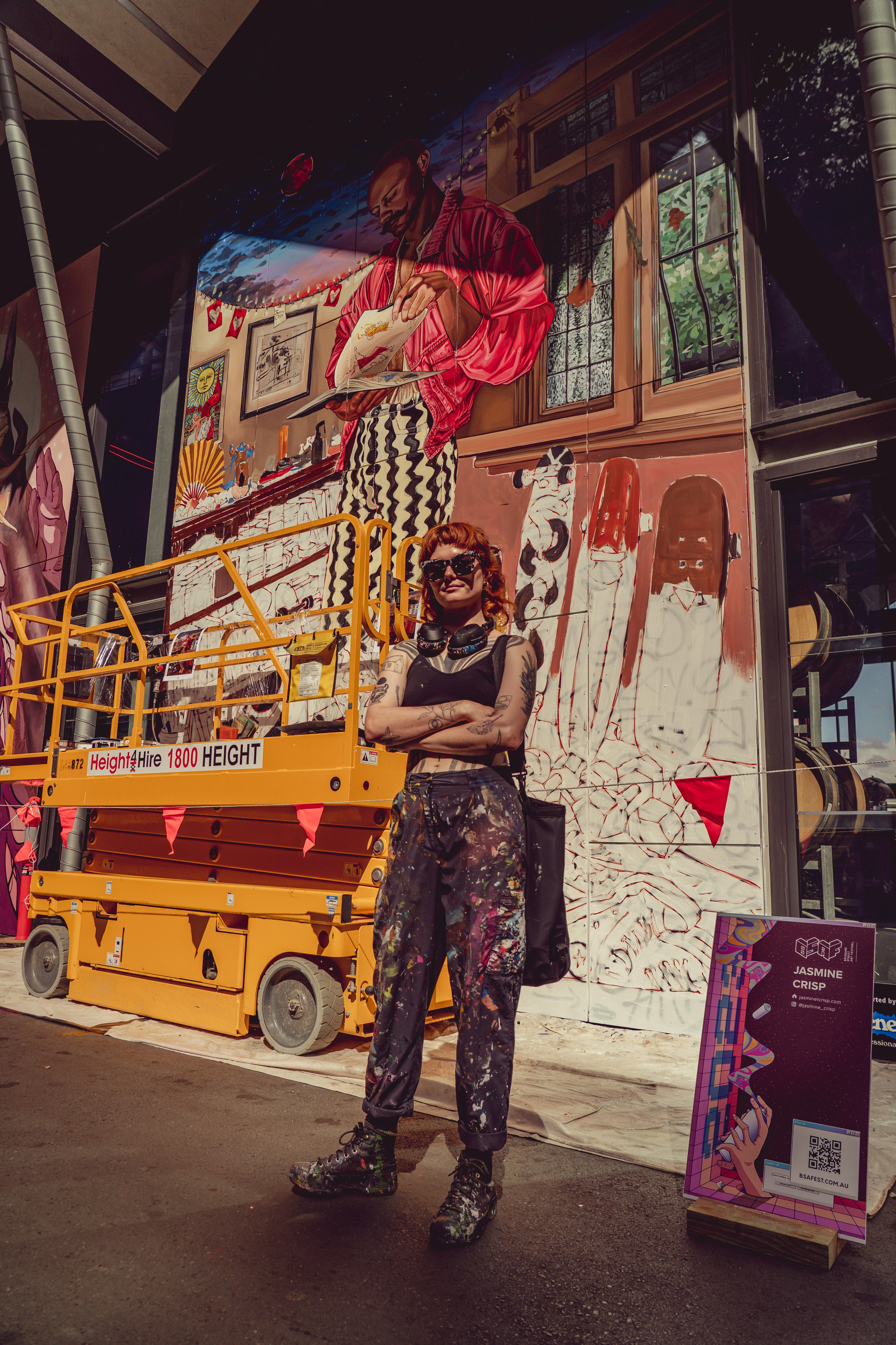 Strong photo of the artist standing in front of an artwork - their arms are closed and a crane and mural are in the background