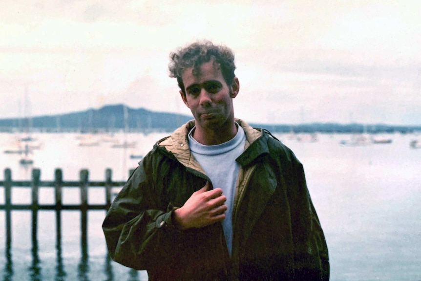 In an old photo Richard Aedy, aged in his 20s, stands in front of a yacht-filled harbour wearing a jacket.
