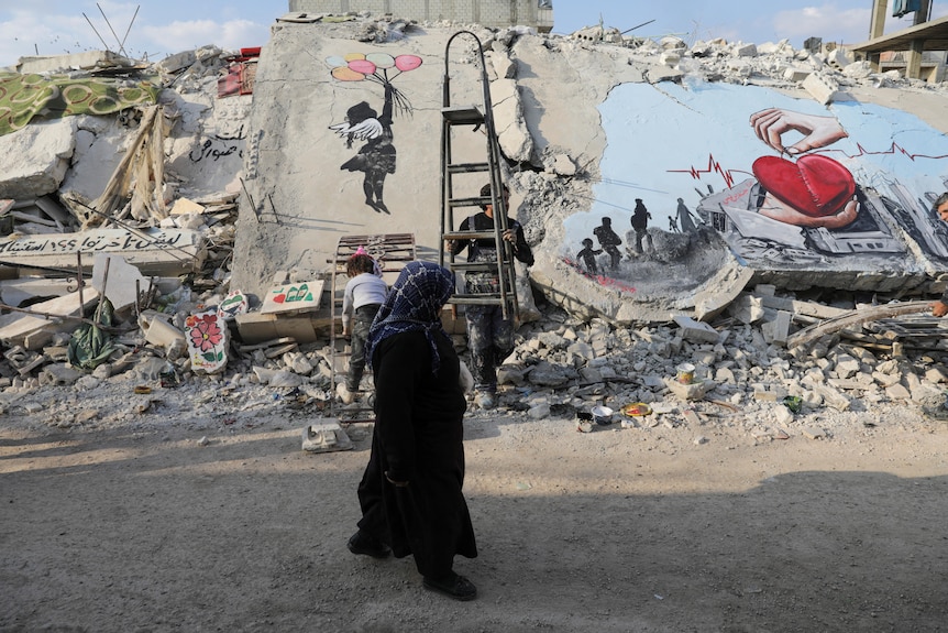 A woman walks past a destroyed building which has a mural of a heart on it. 