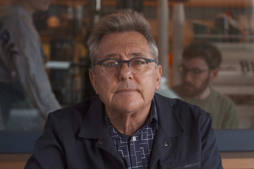 a man with glasses smiling in front of a cafe