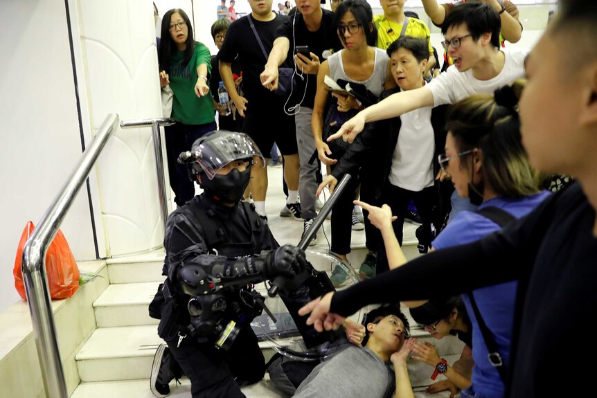 A crowd of people surround a riot police officer and point at him as he detains a protester lying flat against steps.