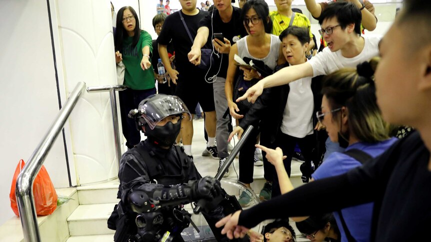 A crowd of people surround a riot police officer and point at him as he detains a protester lying flat against steps.