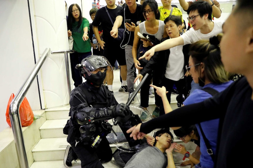 A crowd of people surround a riot police officer and point at him as he detains a protester lying flat against steps.