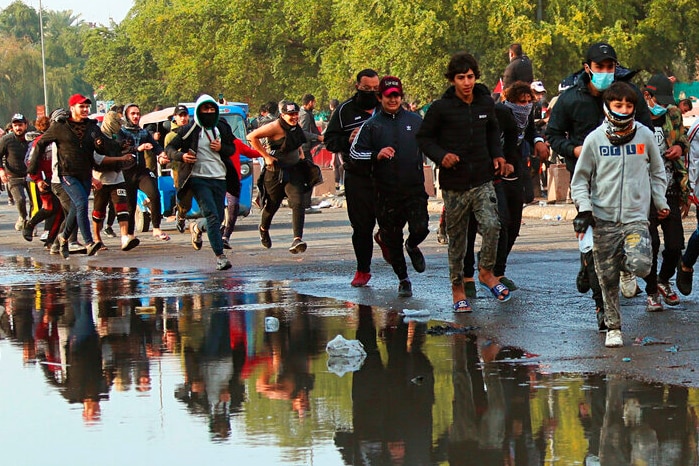 Beyond a pool of rainwater, you see a group of young protesters running, with their reflections appearing in the pool.