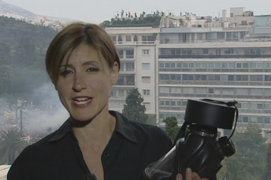 Emma Alberici, a reporter with short brown hair, stands on a balcony holding a gas mask while riots go on behind her.