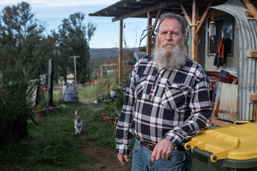 A man with a grey beard outside a shed and leaning on a wheelie bin