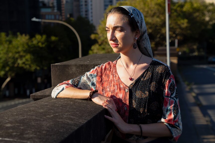 Woman, wearing patterned blouse and hair pulled back by loose headscarf, leaning on cement and looking towards sun.