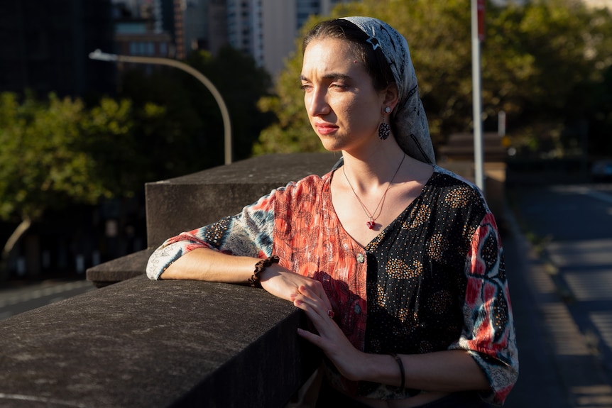 Woman, wearing patterned blouse and hair pulled back by loose headscarf, leaning on cement and looking towards sun.