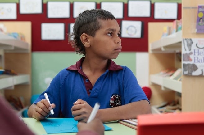 An Aboriginal boy at a school desk in uniform writing on a piece of paper