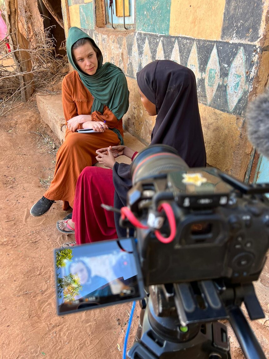 Woman in a scarf interviewing another woman in a scarf as camera films them.