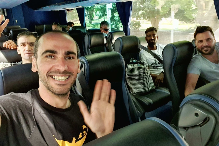 A man on a bus takes a selfie, with him and others in the seats around him all smiling happily.