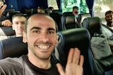 A man on a bus takes a selfie, with him and others in the seats around him all smiling happily.