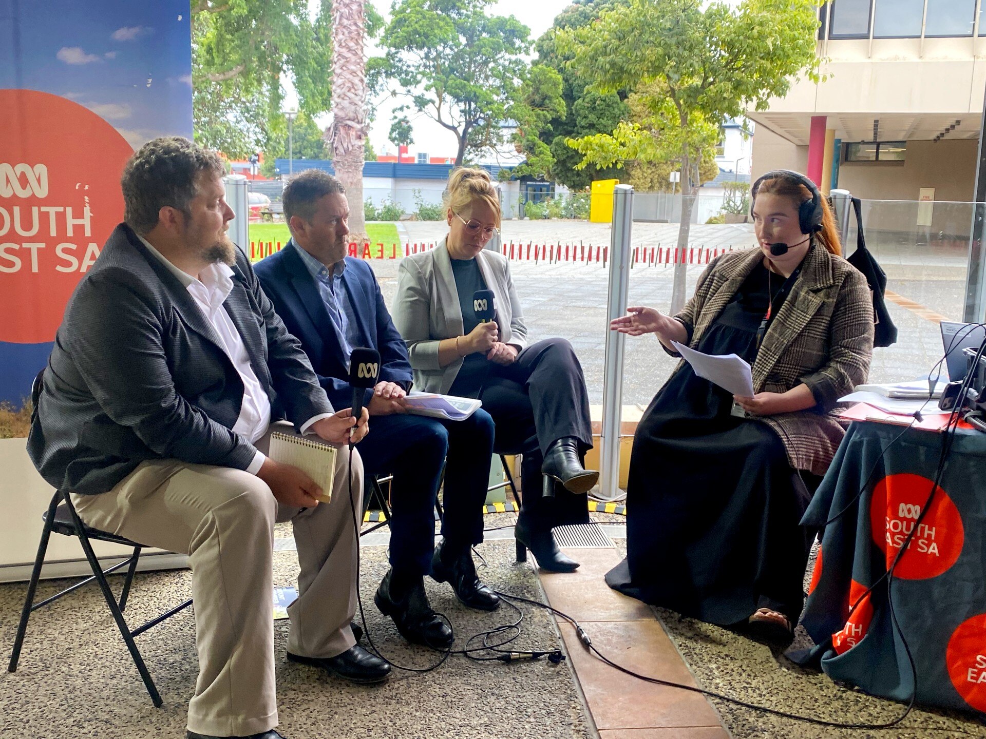 Three people sit down with a mic with a radio presenter holding a sheet with ABC in background
