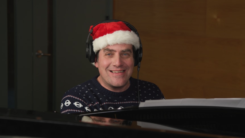 Russell Torrance sitting behind a piano, wearing a Christmas jumper, a santa hat and headphones