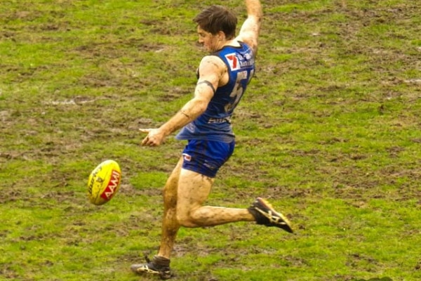 A muddy football player in a blue and black guernsey kicks a ball on a field.