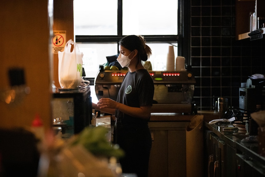 A woman in a mask stands behind a restaurant counter.