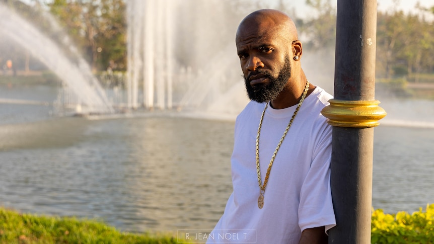 DJ Just Dizle standing outside, leaning on a pole, in front of a river, wearing a white t-shirt and gold chain