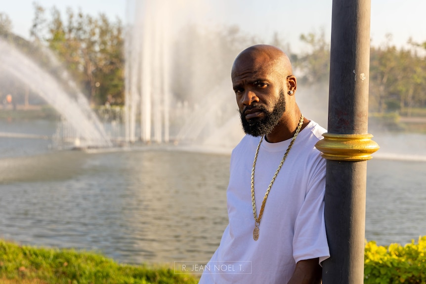 DJ Just Dizle standing outside, leaning on a pole, in front of a river, wearing a white t-shirt and gold chain