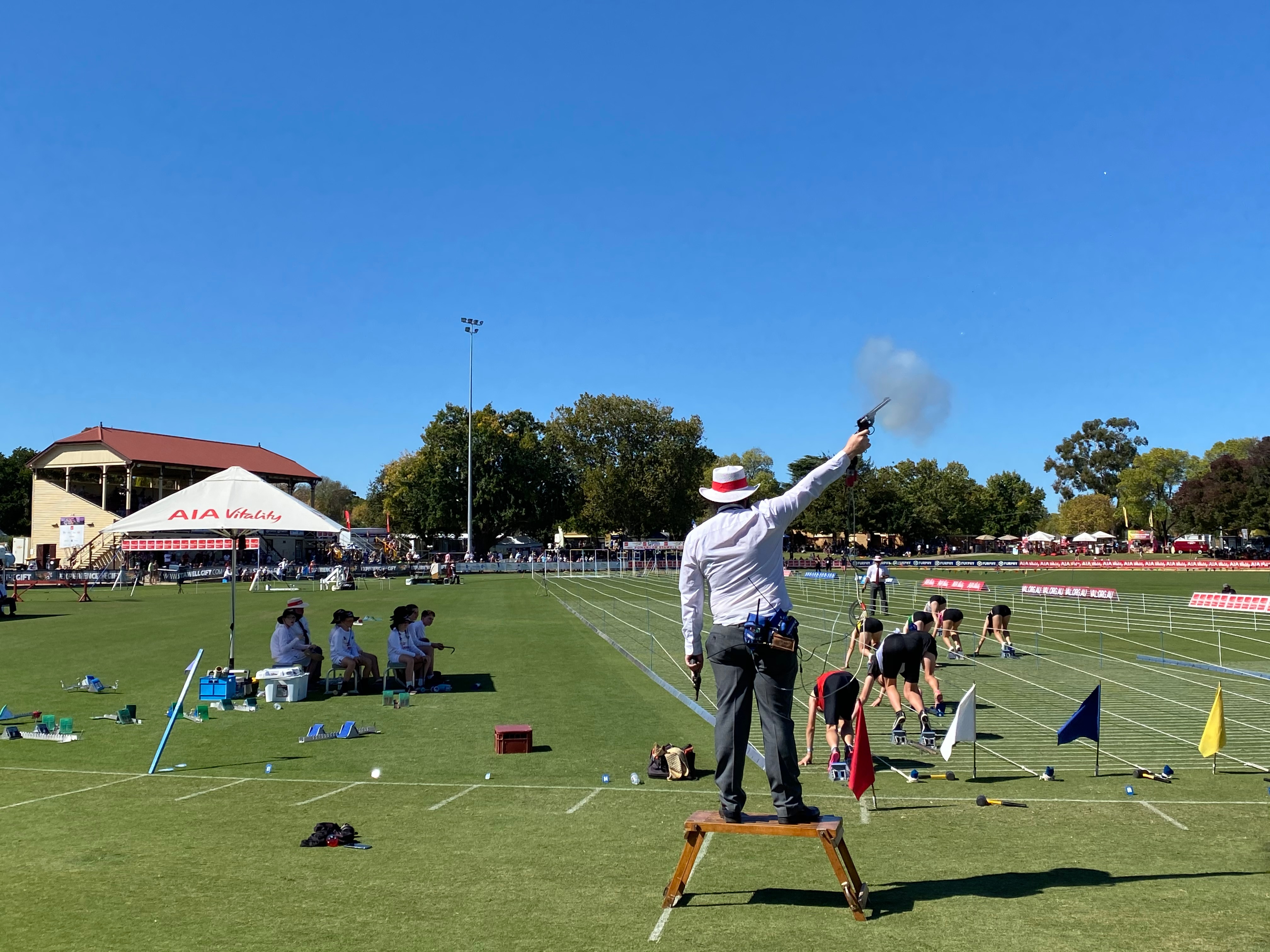 A man stands on a box and fires a gun. Meanwhile, several young children on their marks prepare to run towards a grandstand