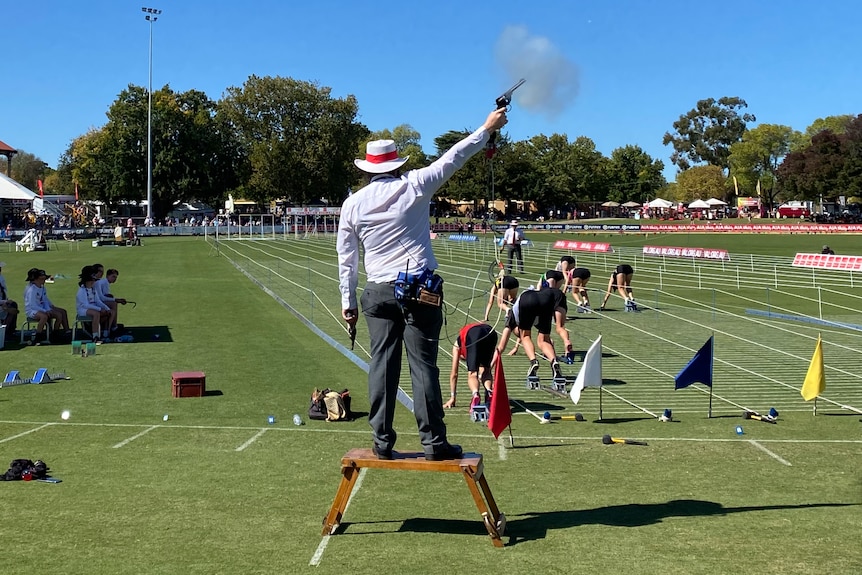 A man stands on a box and fires a gun. Meanwhile, several young children on their marks prepare to run towards a grandstand