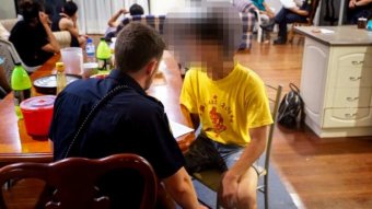 Man wearing ABF uniform interviews man, pixelated, wearing yellow t-shirt in a house with several other people of Asian descent.