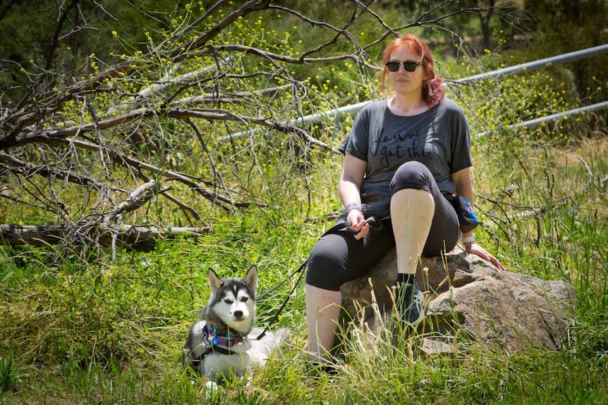 Janine Monty sits with her dog on a large rock surrounded by parklands.