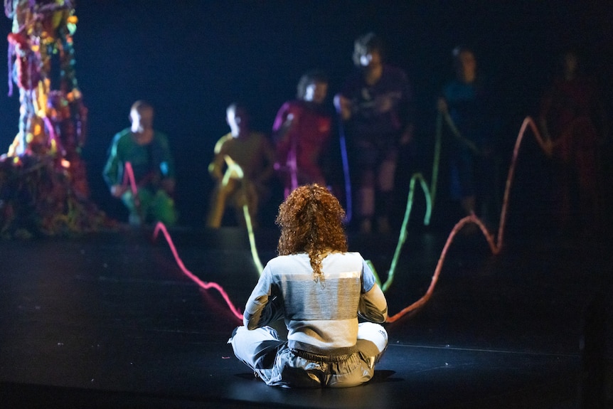 Uma dançarina se senta no palco segurando cinco longos fios coloridos de lã