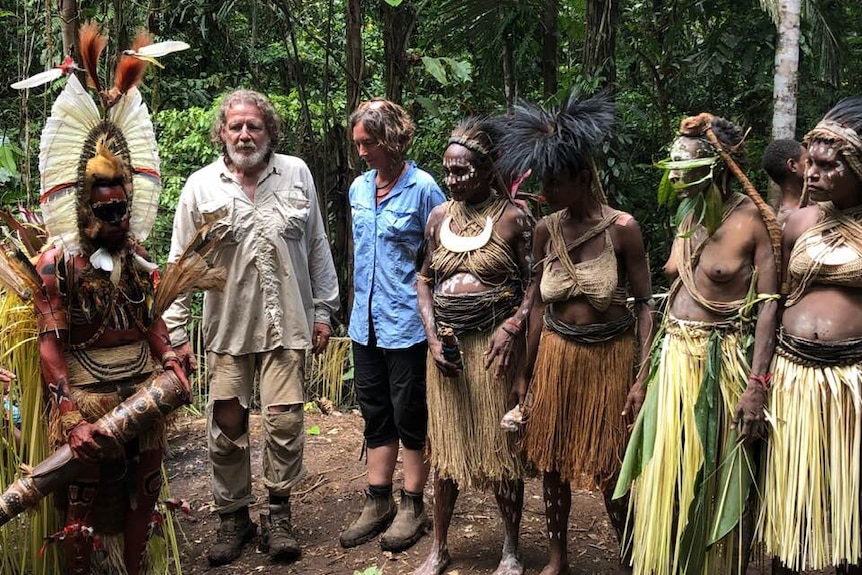 A man and woman in hiking clothes stand with a group of people in traditional PNG dress