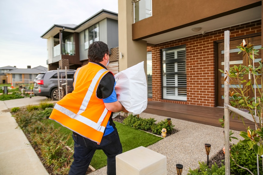 A man in an orange high-vis vest is photographed from behind carrying a large white package while walking towards a house.