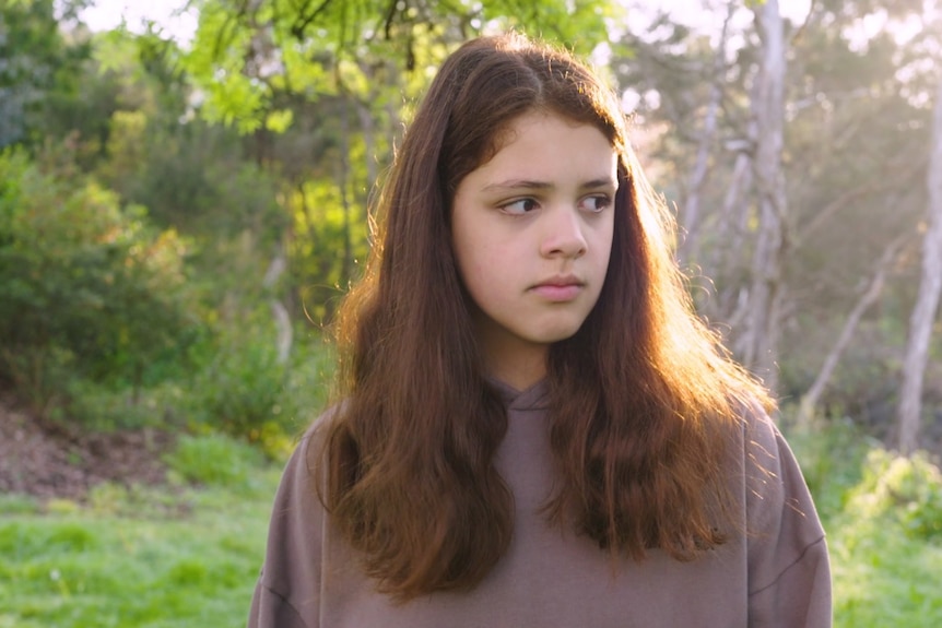 A young teenage girl with long dark hair wearing a brown jumper looks to her left while walking through a park.