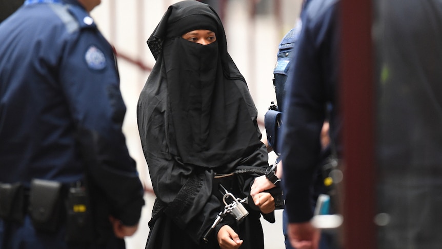 A woman wearing a black niqab and black robes wearing handcuffs surrounded by police.