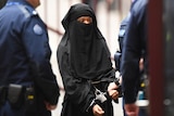 A woman wearing a black niqab and black robes wearing handcuffs surrounded by police.