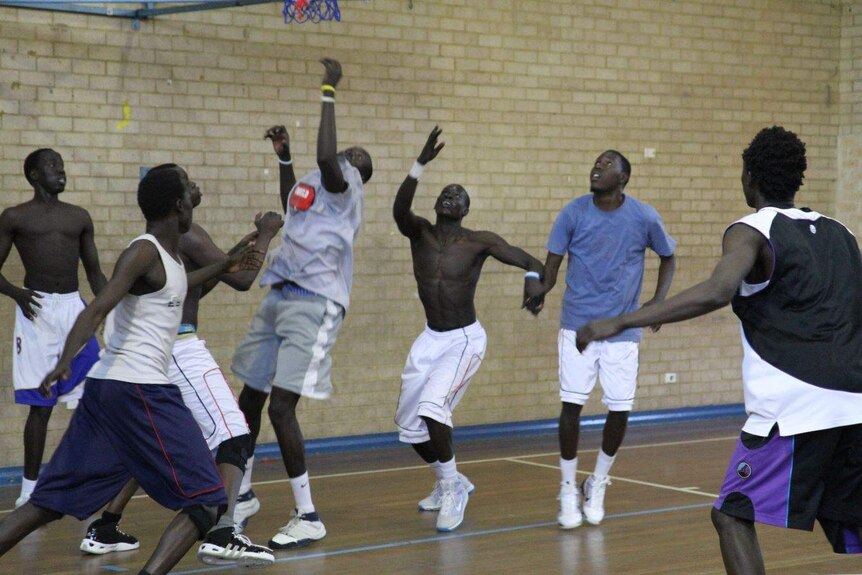 A group of seven men play basketball in a brick building, some with shirts off