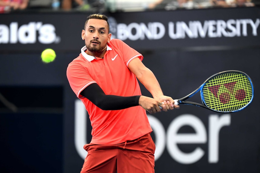Nick Kyrgios wearing a pale red shirt and one black arm warmer prepares to hit the ball with both his hands on his racquet.
