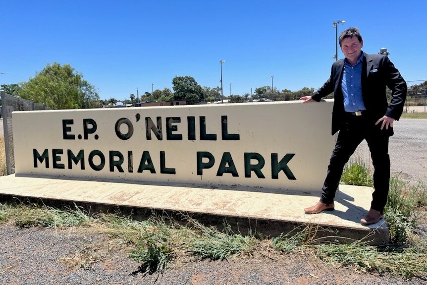 A smiling, dark haired man dressed in a dark suit and squinting in the sun. He is leaning on a sign for a public park.