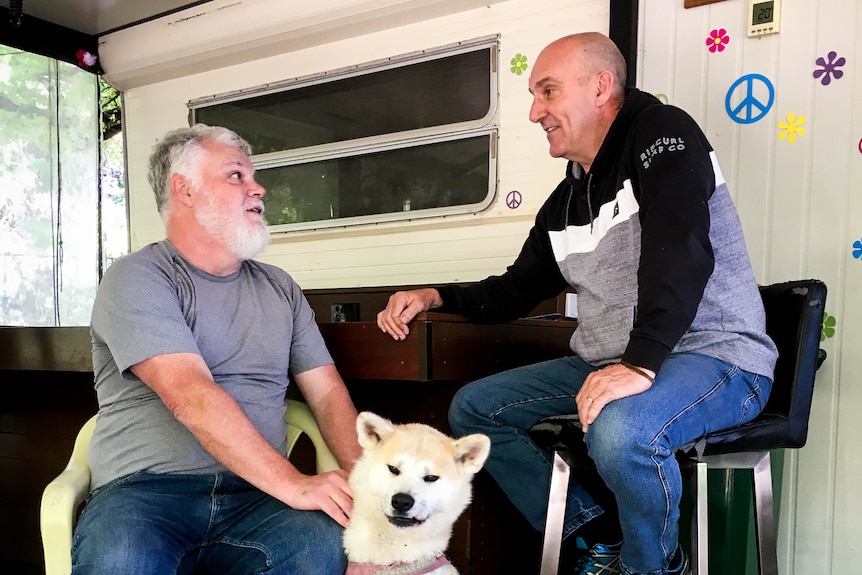 two men chat with a dog at their feet in the annex of a caravan.