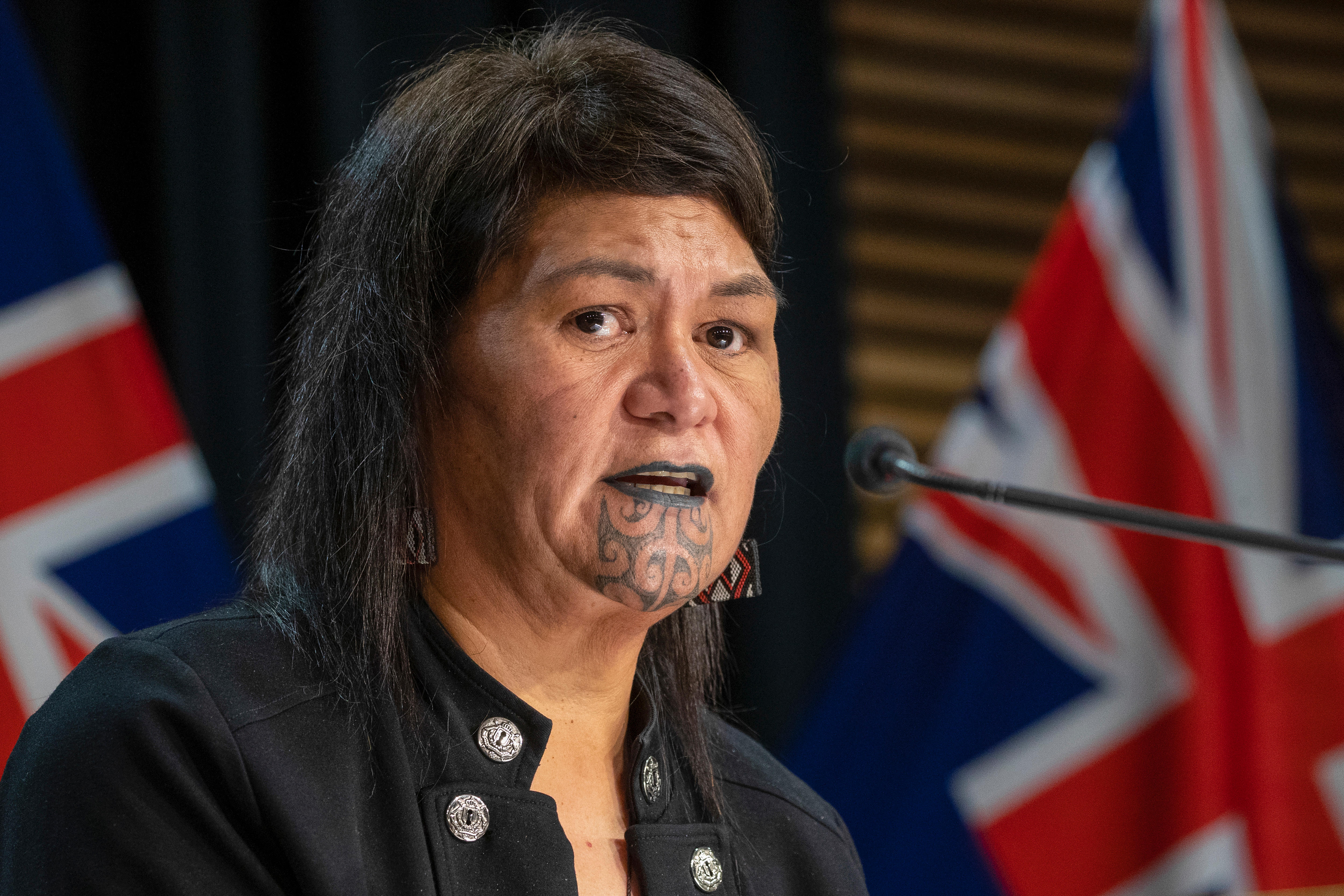 A woman with a traditional chin tattoo speaks into a thin black mic rigged to a lecturn, with the Union Jack seen behind