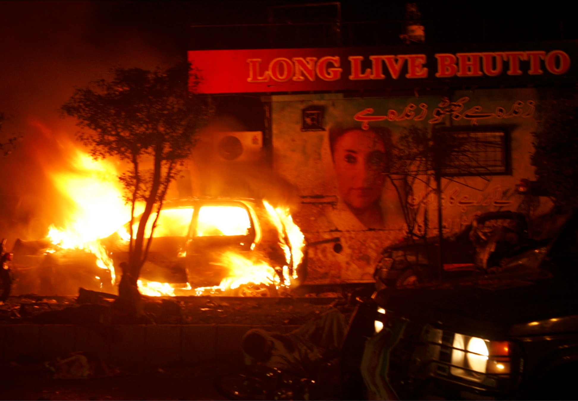 Um carro pega fogo sob uma placa que diz:" VIVA BUTTO" com uma foto dela.