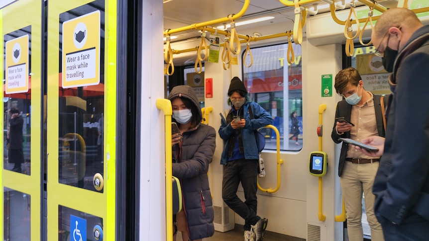 Four commuters wearing masks on a Melbourne train who are looking at their phones through the open door.