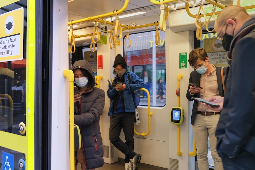 Four commuters wearing masks on a Melbourne train who are looking at their phones through the open door.