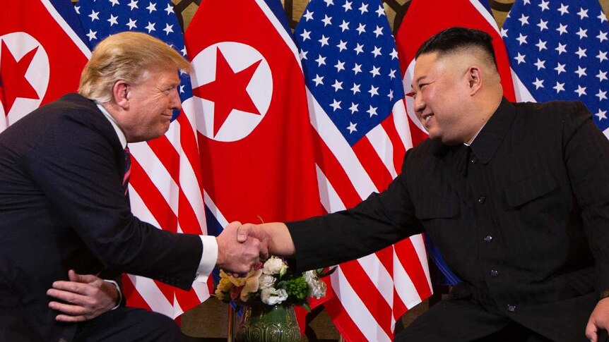 Donald Trump and Kim Jong-un shake hands while they sit in chairs in front North Korean and United States flags.